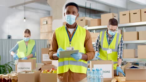 front view of african american male volunteer in facial mask checking donation list and smiling to the camera in warehouse house