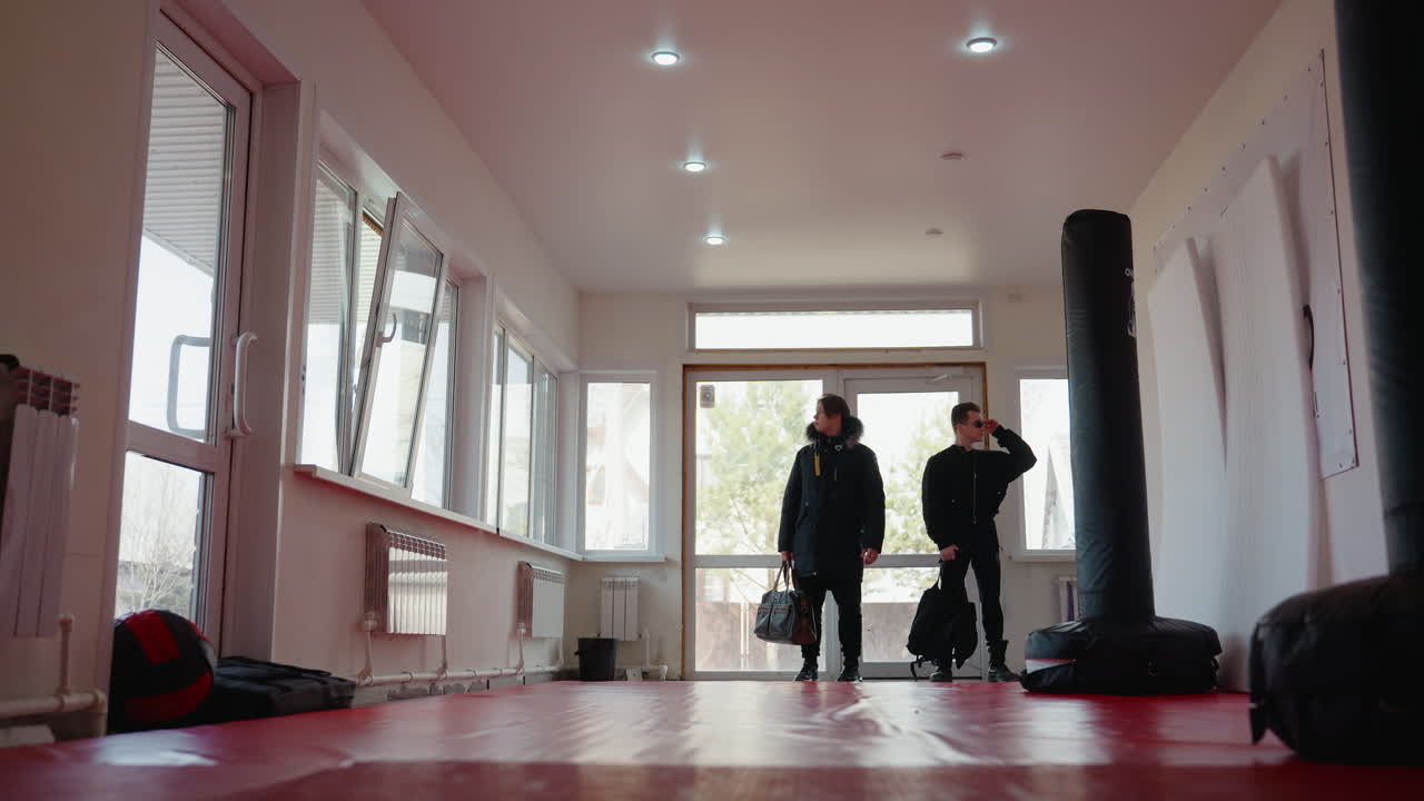 Two guys in black jackets walk into bright training hall through glass door, carrying bags, entering gym with punching bags, red mats, large windows and modern lighting, representing active lifestyle scene