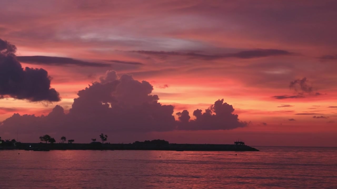 Static medium tracking shot panning right across silhouetted coastal land and clouds illuminated by a vibrant pink and orange sunset over Tanjung Tokong sea.