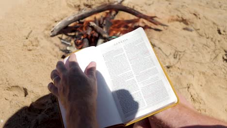 a close shot slow motion shot of a man opening up a bible besides a fire on the beach