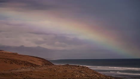 lapso de tiempo de un enorme arco iris sobre la costa rocosa costera mientras las olas rompen la costa lunar