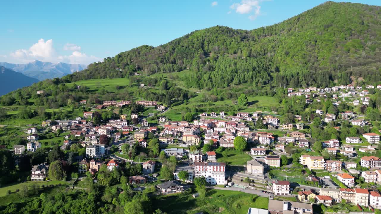 Sideways over San Fedele near Como, the view reveals charming rooftops and narrow streets nestled between vibrant hills and imposing mountains, all bathed in warm summer light as captured by a drone