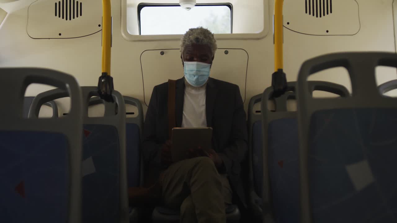 hombre mayor afroamericano con máscara facial usando una tableta digital mientras está sentado en el autobús