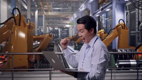 side view of an asian male professional worker standing with his laptop with industrial robotic arms working, he raises his fist up with screaming goal after typed on the laptop