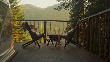 sister and brother sit on sunny terrace engaged in playing video game. little boy and girl relish pleasant weekend morning against awesome view of mountains