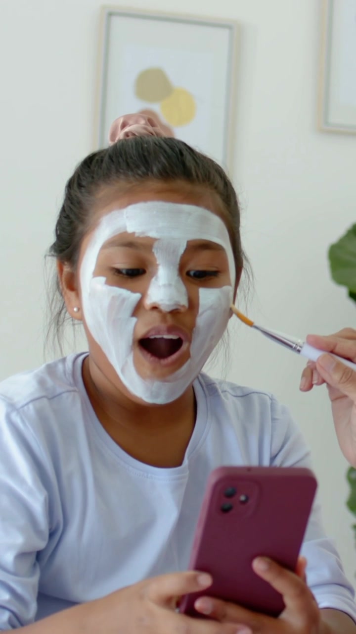 Video vertical: Adolescente sosteniendo un teléfono y reaccionando mientras una mano aplica una mascarilla con un pincel para el cuidado de la piel en casa.