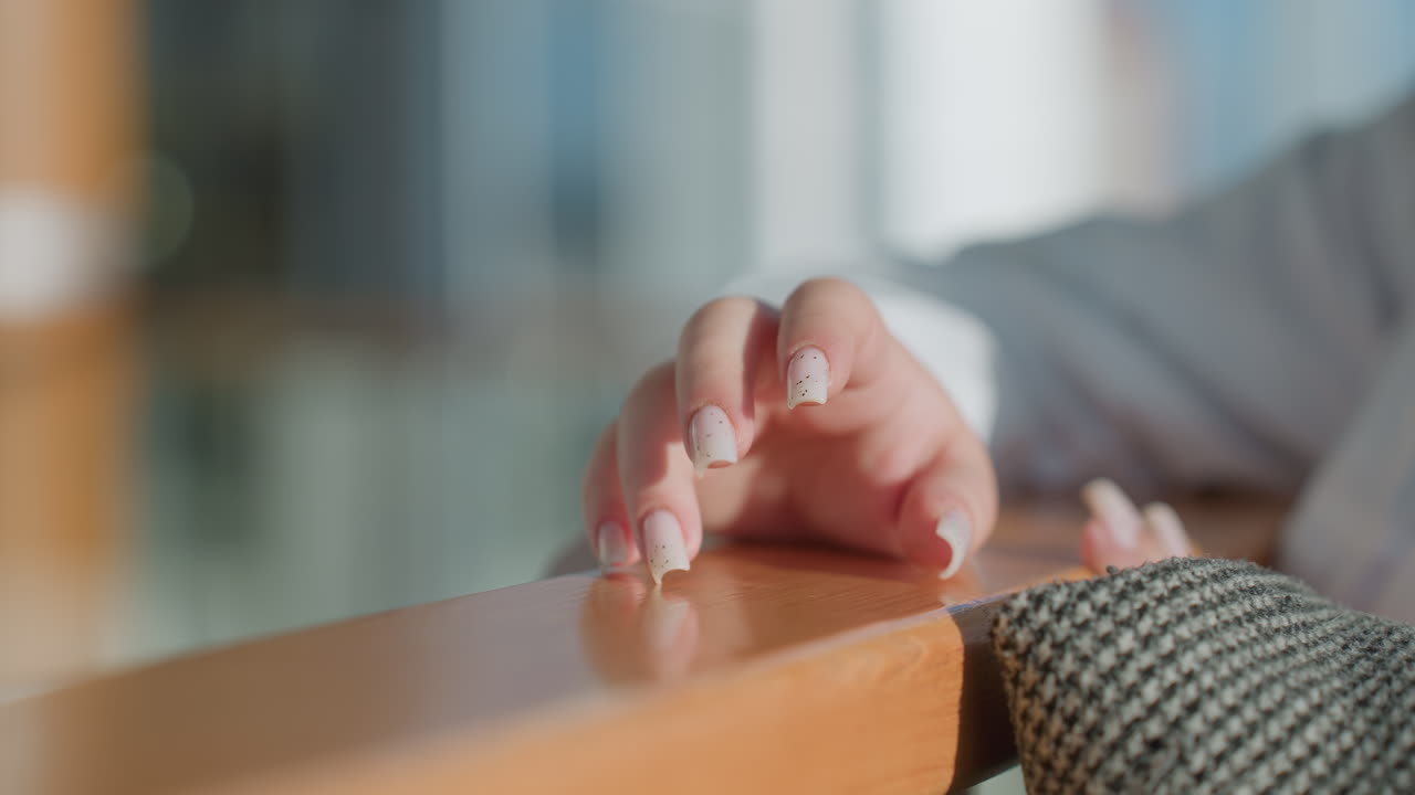 Close up of fair skinned hand with manicured nails gently tapping on polished wooden rail in modern mall interior with sunlight reflections and soft blurred background