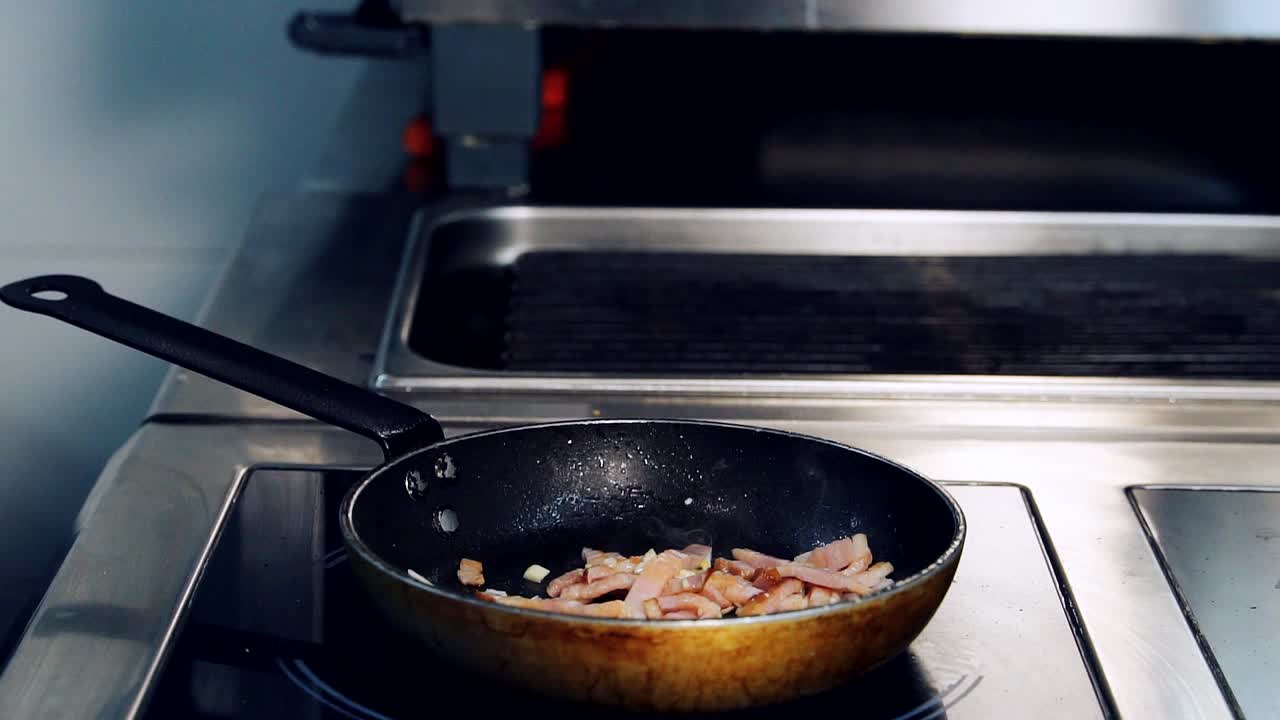 Frying pan with meat on a hot stove. Professional cook turning over pieces of bacon frying in the restaurant kitchen. Close-up