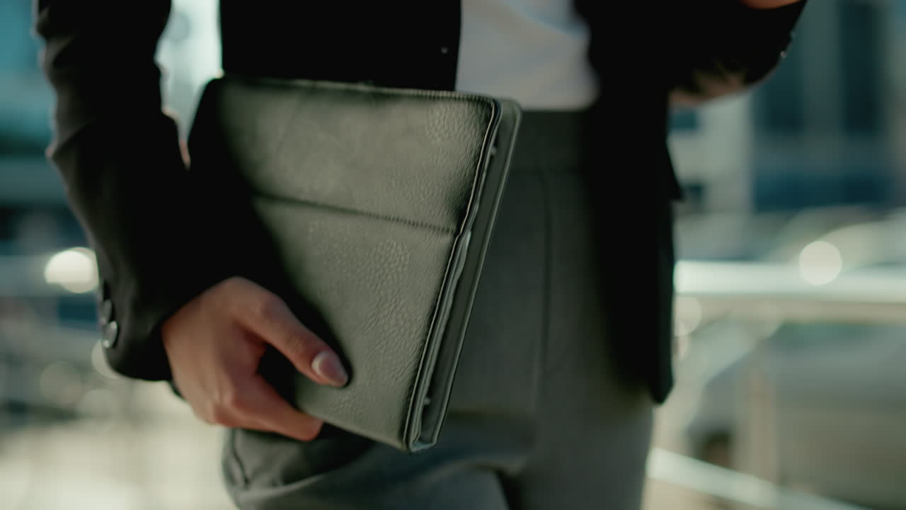 Close up partial view of professional woman holding tablet and coffee cup walking beside iron railing as she sips drink with urban background and parked cars softly blurred by sunlight reflection