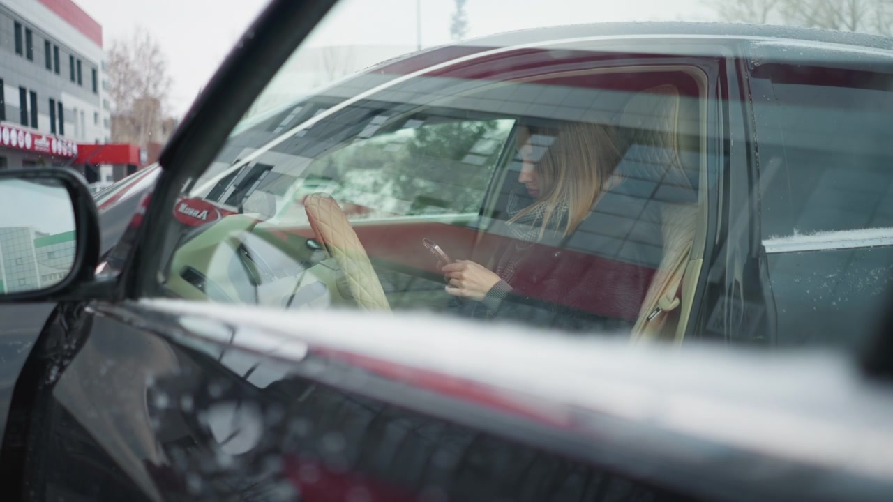 Close up of woman sitting in car holding phone while driving. Car interior with steering wheel and dashboard visible. Out of focus urban background through windshield