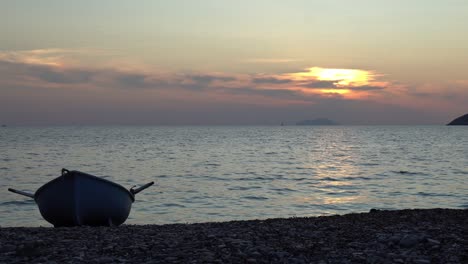 un bote de remos solitario se sienta en una playa de guijarros mientras el sol se pone detrás de él y las suaves olas llegan