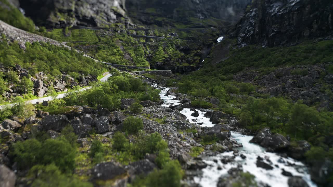 A rapid stream flows through a dense green valley, cascading down the mountain beneath the Trollstigen road as cars and motorhomes climb the road slowly. A tilt-shift video.