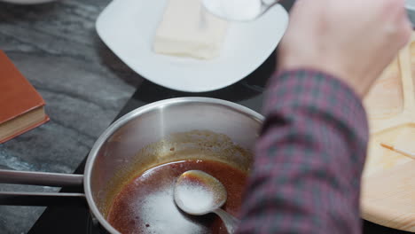 close-up of cook pouring salt into stew while stirring with a spoon inside pot, various kitchen utensils, including a wooden board and a cookbook, are arranged nearby