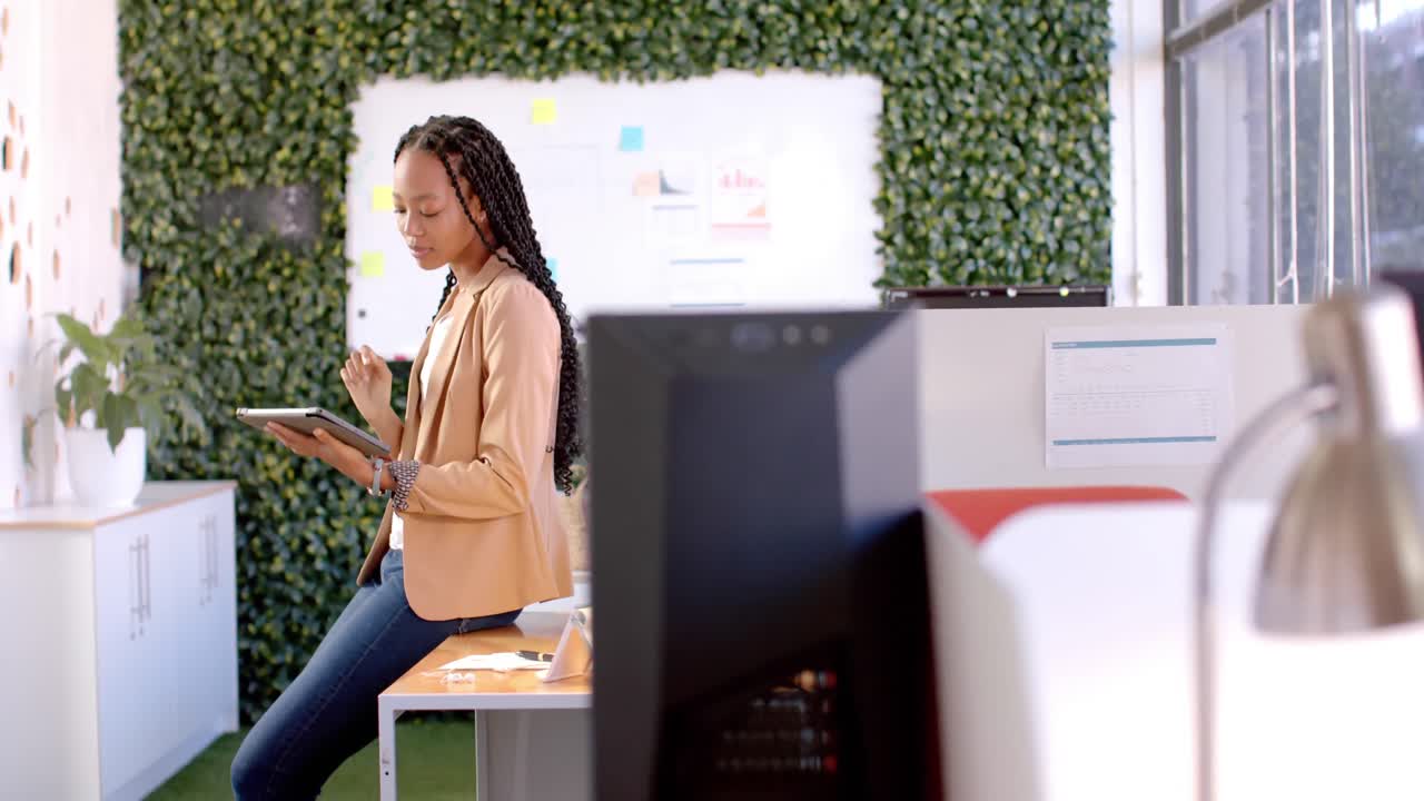 Happy african american casual businesswoman sitting on desk using tablet in office, slow motion