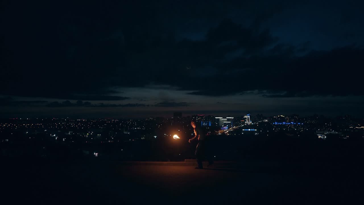 Giovane uomo biondo esegue trucchi con il fuoco, sputa fuoco di notte con lo skyline della città sullo sfondo.