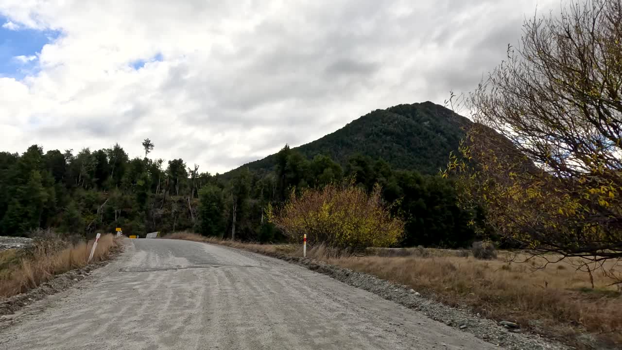 Vehicle travels gravel road through mountainous landscape, approaching bridge under cloudy daylight, steady forward motion