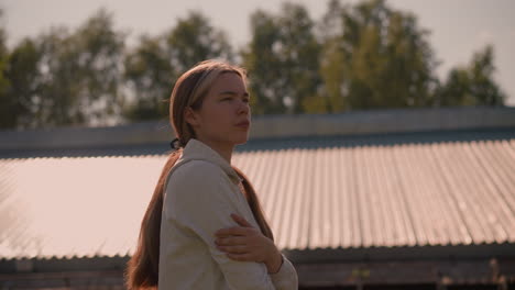 young girl with long hair tied back in hoodie stands outdoors, looking pensive with thoughtful expression, sunlight casts a warm glow on her, with roof and trees in background