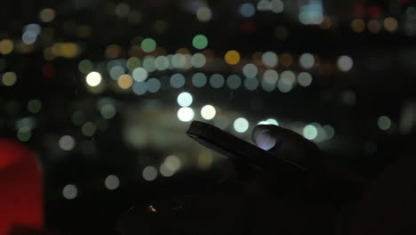 Navegando-En-El-Teléfono-Inteligente-Por-La-Noche-Las-Luces-De-La-Ciudad-En-Segundo-Plano.