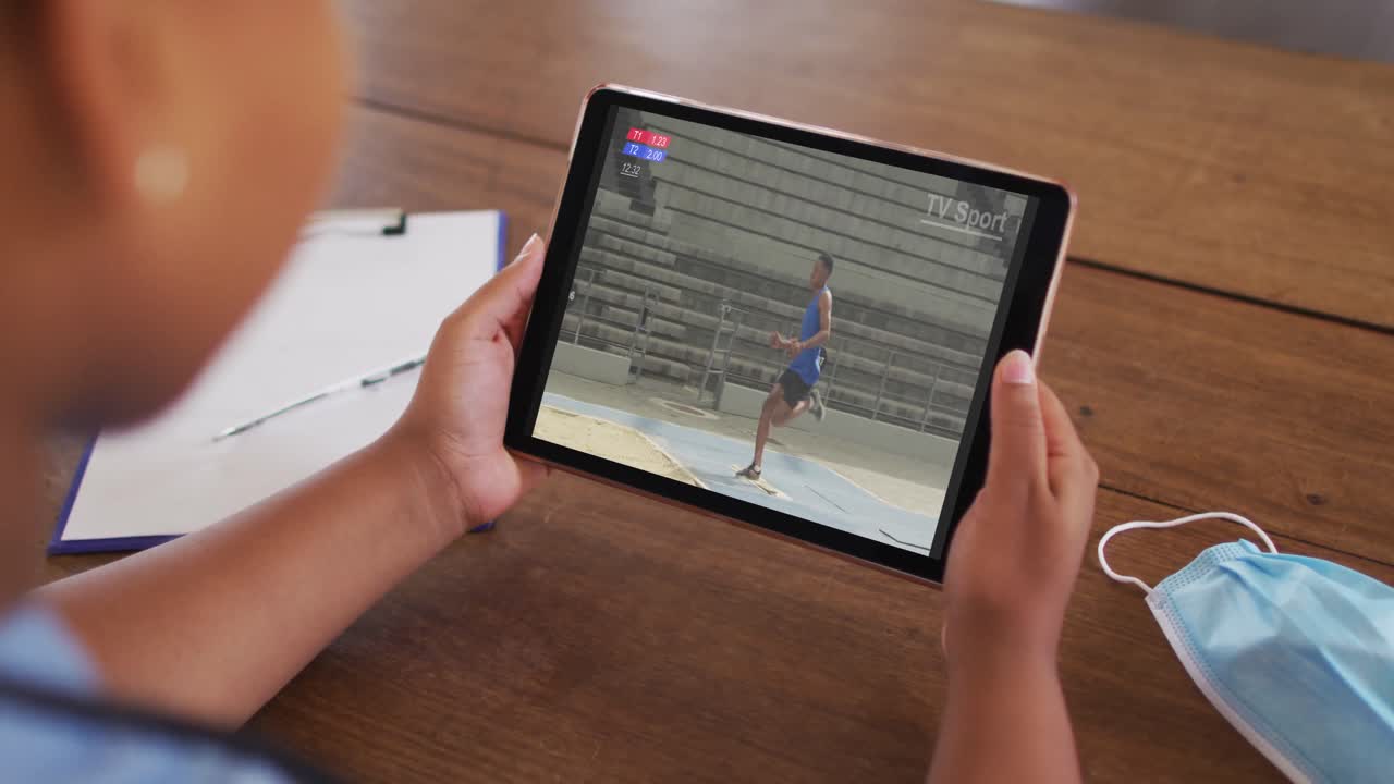compuesto de una mujer sentada en una mesa con una máscara facial, viendo el evento de salto de longitud de atletismo en la tableta