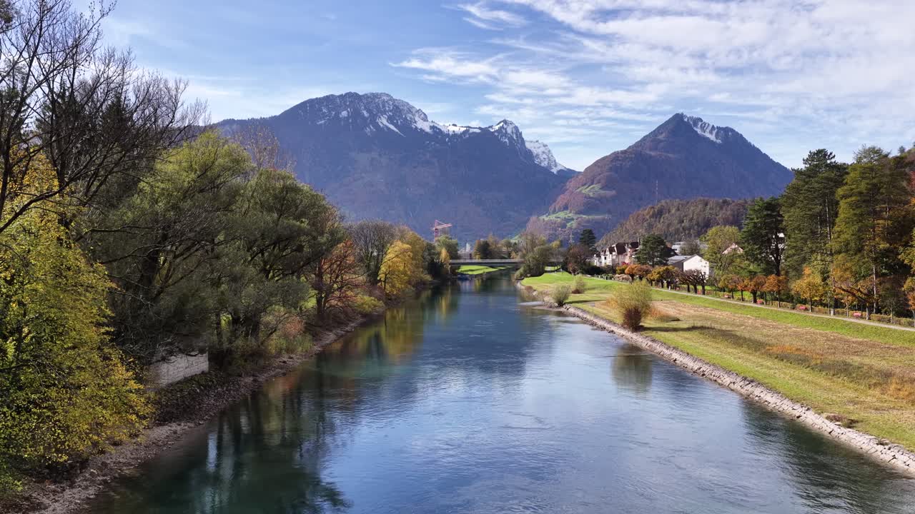 The Linth River near Weesen, Switzerland, flows between tree-lined banks with autumn color, leading toward snow-capped alpine mountains. Nature, travel, and environmental footage for various uses