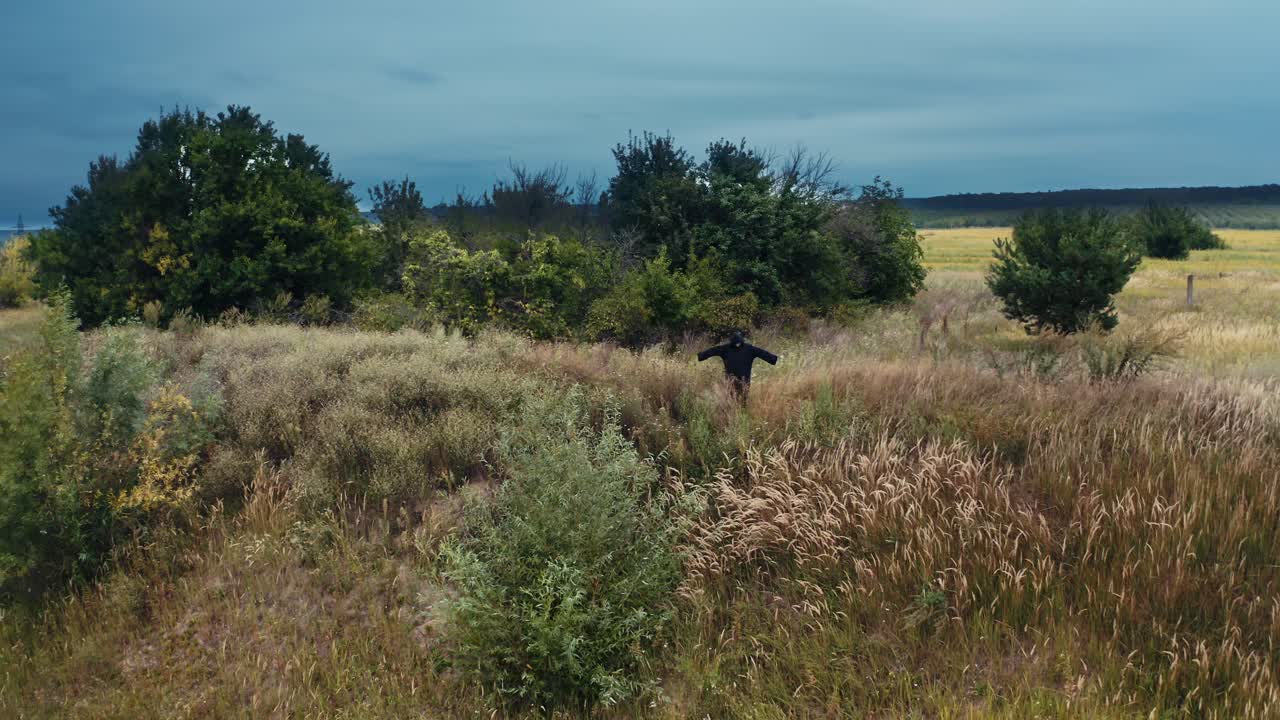 Devil in black costume in nature. Field in summer and dark figure in black robe with outstretched arms standing. Horrorful ghost outdoors.