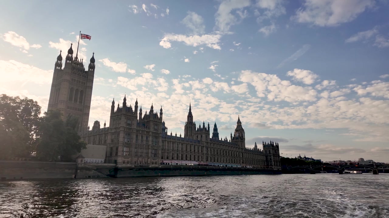 Westminsterpalasset strekker seg langs vannet mens en båt glir forbi under en varm himmel i den gylne timen.