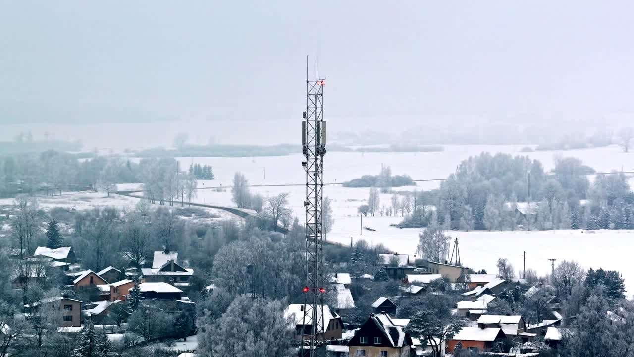 Drone view circling tall telecommunication tower and cluster of houses nestled among frosted trees and white fields in quiet hazy winter morning in Jelgava Latvia