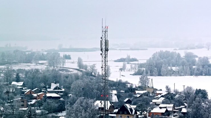 Drone view circling tall telecommunication tower and cluster of houses nestled among frosted trees and white fields in quiet hazy winter morning in Jelgava Latvia