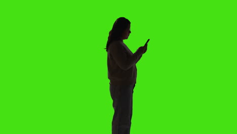 silhouette of woman sending or reading text message or scrolling online on mobile phone standing against green screen with low key lighting