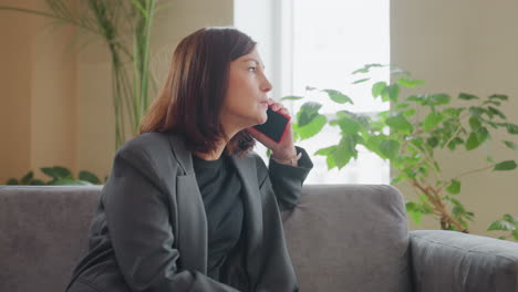 worried businesswoman in gray blazer talking on phone with serious expression while sitting on couch in modern indoor setting surrounded by green plants and natural light through large window