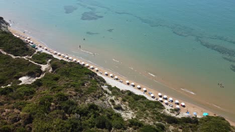 Vista-Aérea-De-Una-Playa-Mediterránea-Con-Un-Mar-Turquesa-Claro,-Bañistas,-Sombrillas-Y-Turistas-Durante-El-Verano-Corfú-Grecia