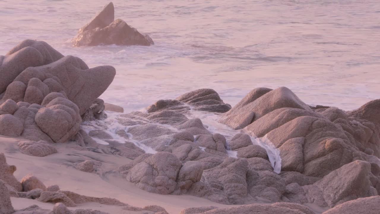 Wide shot of a sandy beach with waves crashing on large rocks, focused on the natural coastal landscape under a soft, pastel sky.