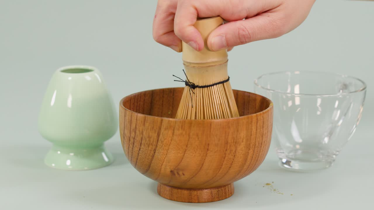Hand uses bamboo whisk to froth matcha in wooden bowl, bright lighting, static overhead shot