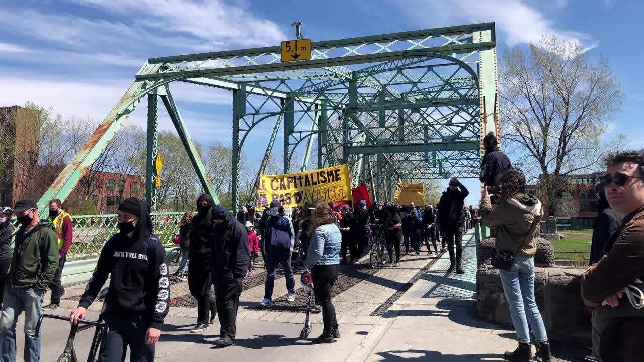 Protestors demonstrating against capitalism, marching across a rusty coloured steel bridge with bystanders filming on their phones.