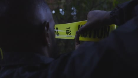 a black police officer hanging caution tape at a crime scene in a wooded area