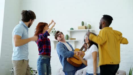 grupo de amigos multiétnicos bailando mientras el joven toca la guitarra y tiene una fiesta en casa en el interior