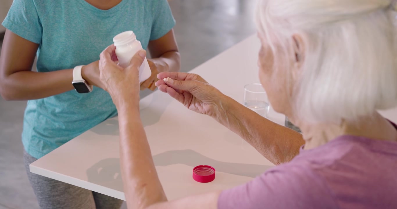 Cuidadora ofreciendo una botella con tapa roja, mujer mayor intercambiando una botella amarilla, preparando medicamentos en la mesa