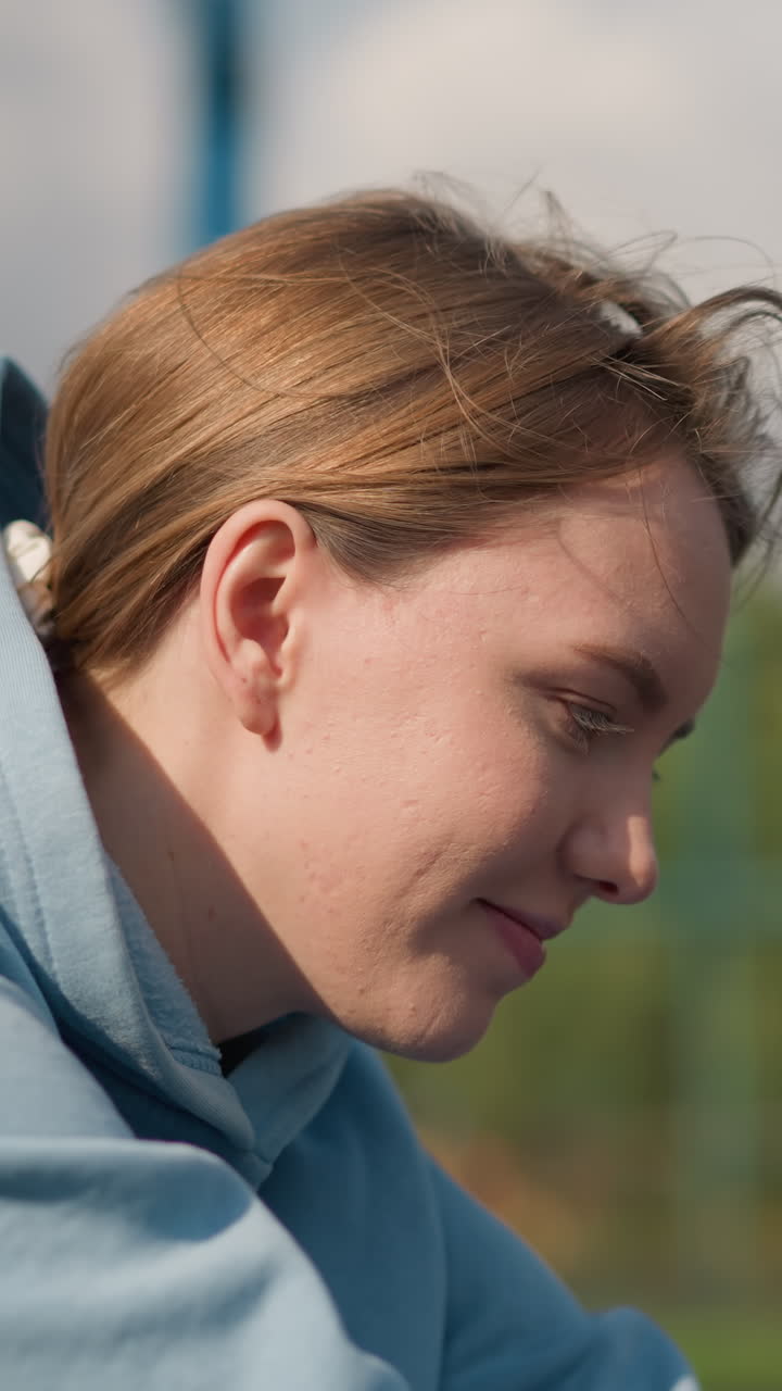 primer plano de una mujer con capucha cian sonriendo cálidamente mientras el viento sopla a través de su cabello con un fondo borroso con cancha deportiva al aire libre y la naturaleza