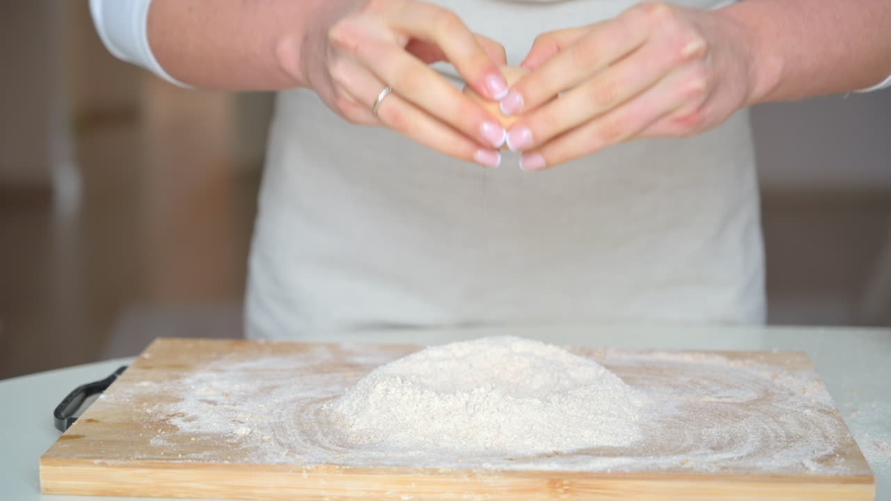 Donna che indossa un grembiule, mentre rompe un uovo nella farina su un tagliere di legno in cucina
