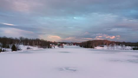 Aerial-footage-flying-fast-and-low-then-rising-up-above-a-frozen-lake-towards-a-small-village-lit-by-the-golden-light-of-sunset