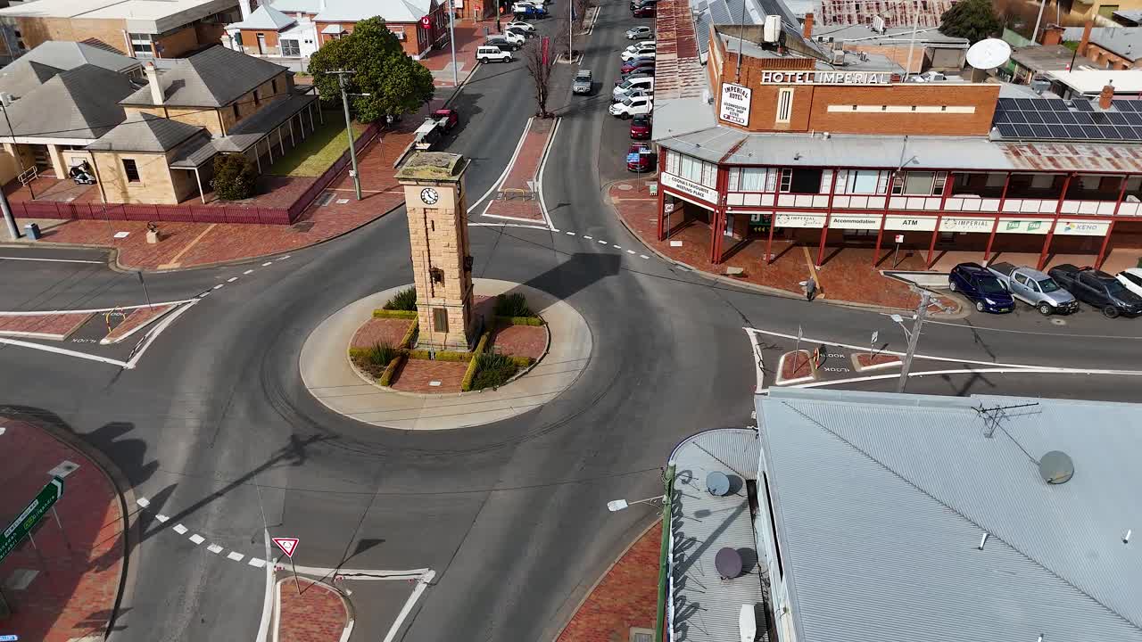 Drone footage shows vehicles navigating a roundabout with a central clock tower in a quiet urban area, under bright daylight with smooth camera movement