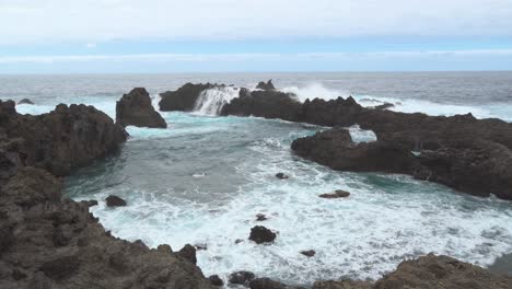 Ocean-Waves-Hitting-The-Black-Rocky-Shoreline,-Sea-Foam,-Blue-Sky,-White-Clouds