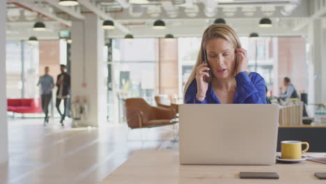 businesswoman sitting at desk on phone call in modern open plan office with colleagues in background