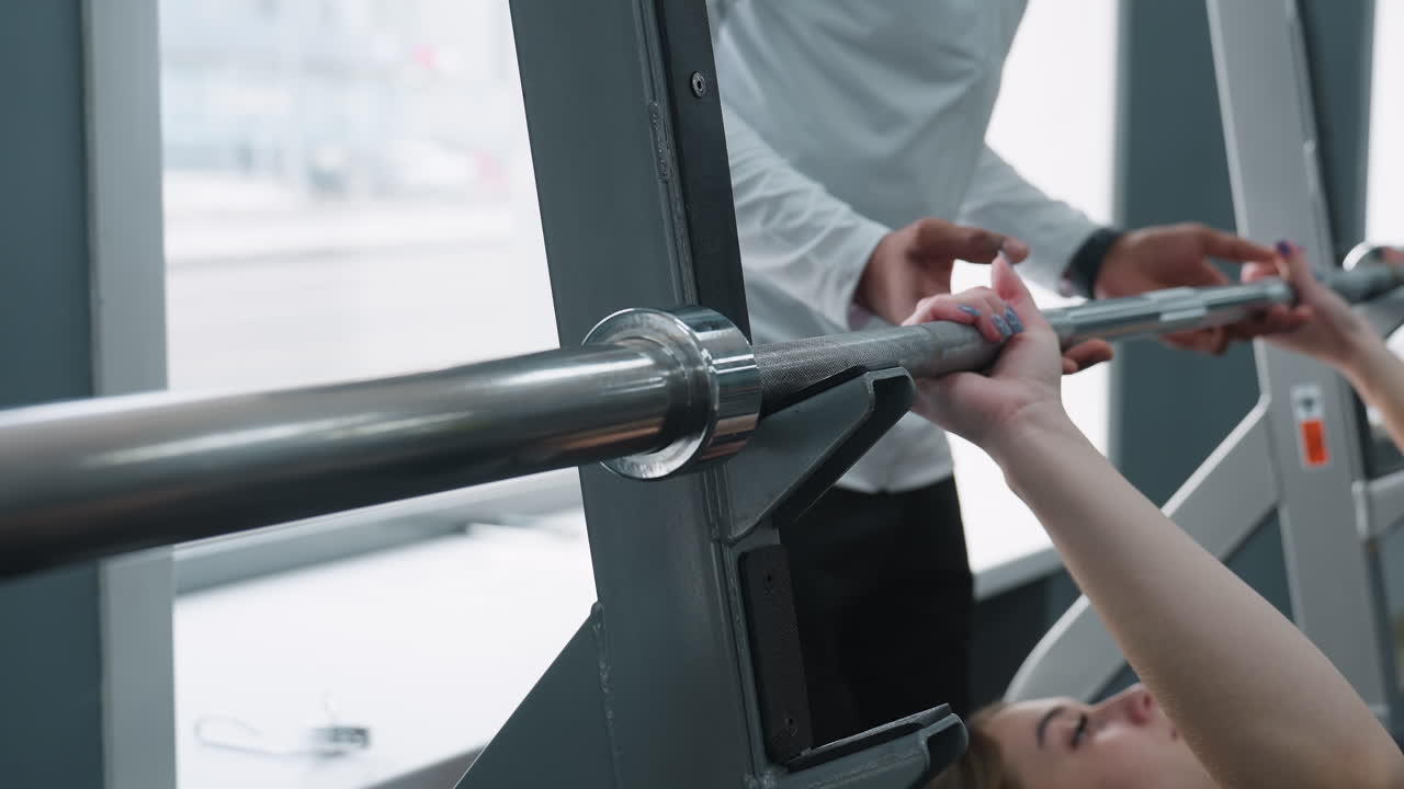 close up of young woman lifting barbell on bench while male trainer assists during workout session in bright gym with large windows, visible city street outside showing passing car and pedestrian
