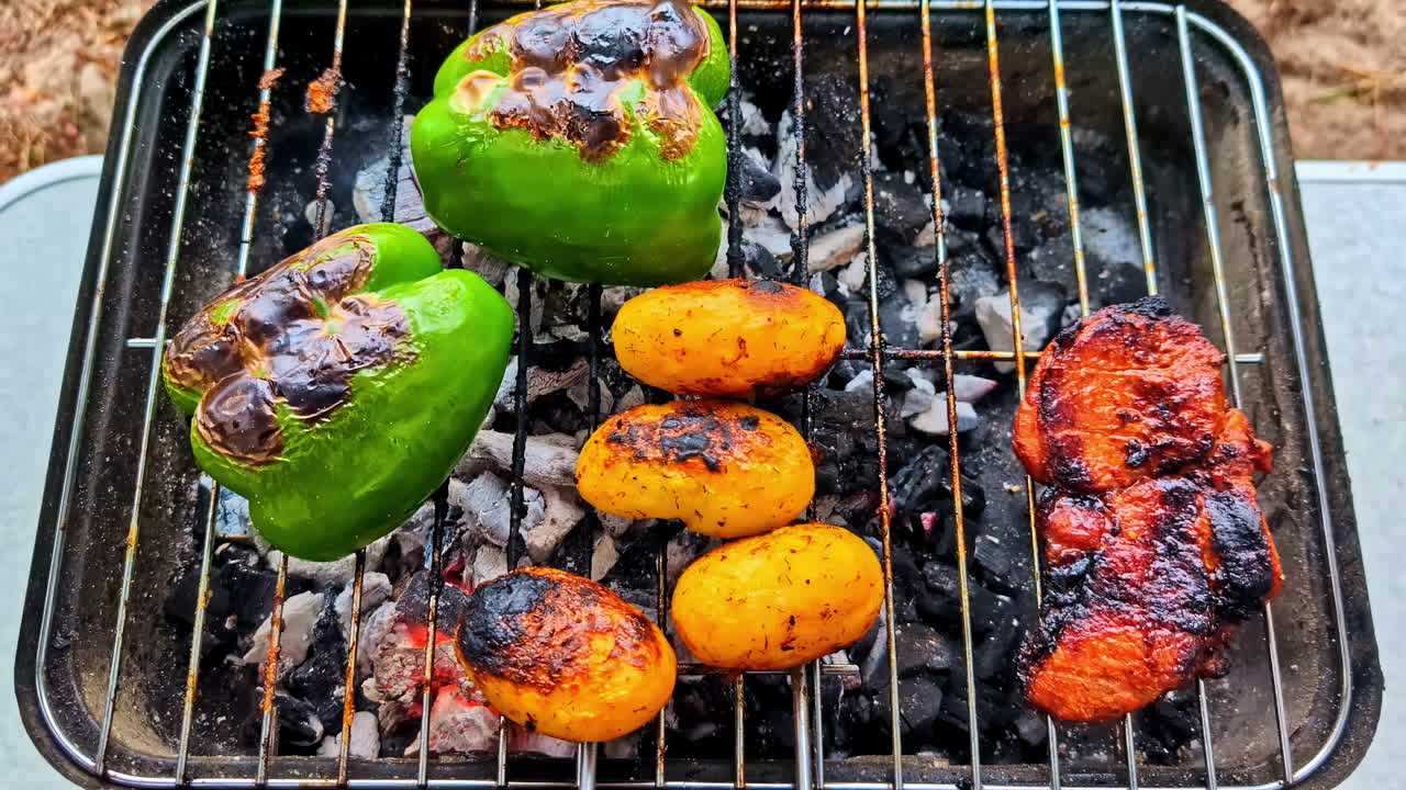A push-in shot moves closer to marinated chicken, green peppers, and potatoes sizzling and charring on a small, portable charcoal grill during an outdoor camp cookout