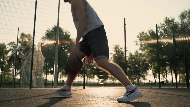 Männlicher Basketballspieler, der den Ball aufprallen lässt, dribbelt und Spin-Moves macht, auf einem Basketballplatz im Freien bei Sonnenuntergang.