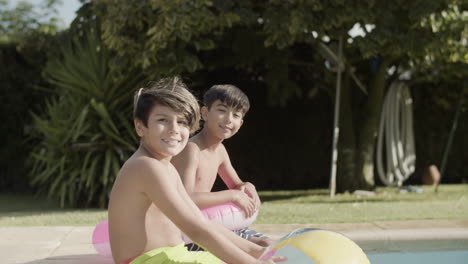 two happy boys sitting on poolside, holding inflatable toys.