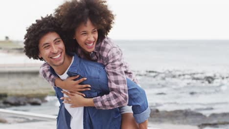 Un-Hombre-Afroamericano-Dando-A-Su-Esposa-Un-Paseo-A-Cuestas-Por-El-Paseo-Marítimo-Cerca-De-La-Playa