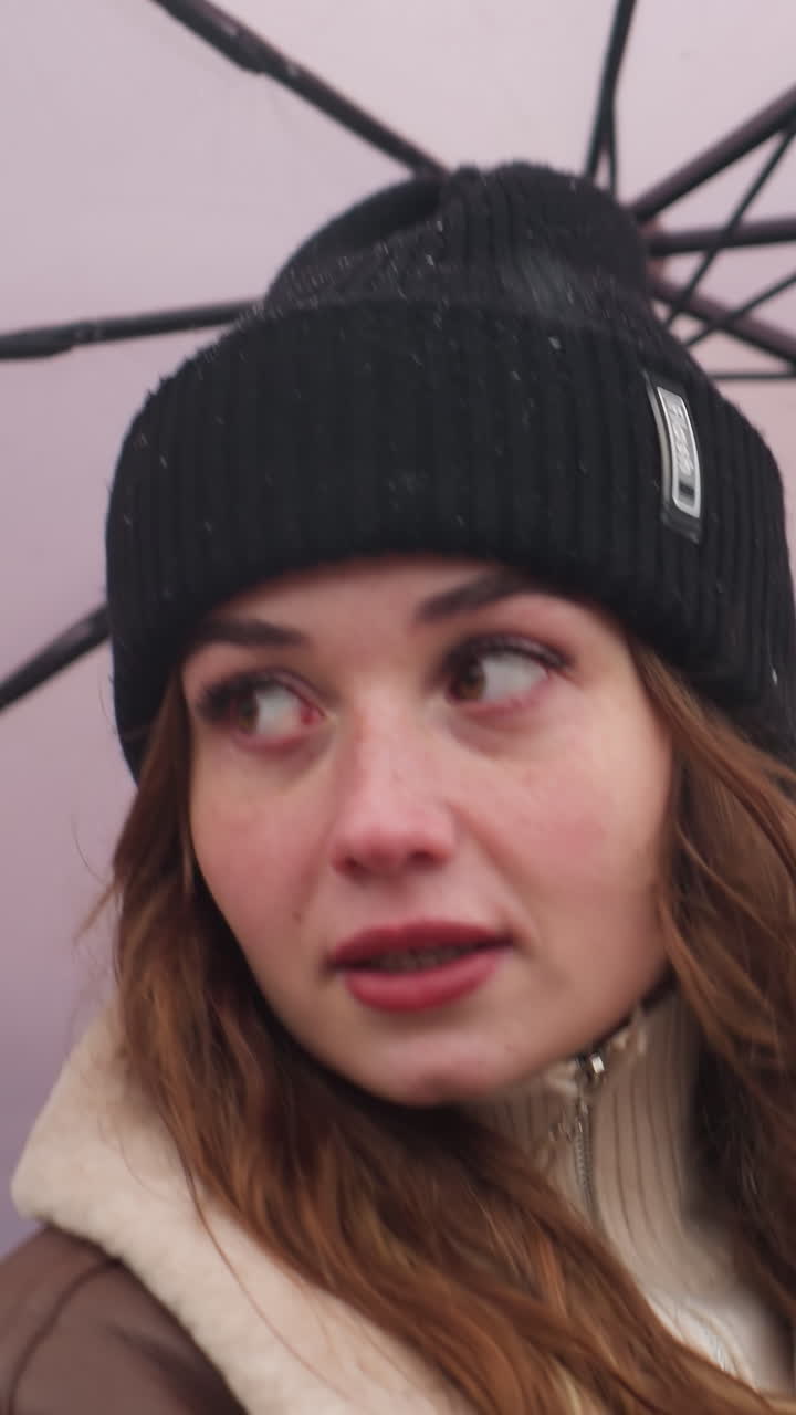 Close shot of happy lady holding umbrella as wind blows gently with light snowfall running outdoors wearing black knit cap and brown shearling jacket strands of hair blowing creating cozy winter vibe