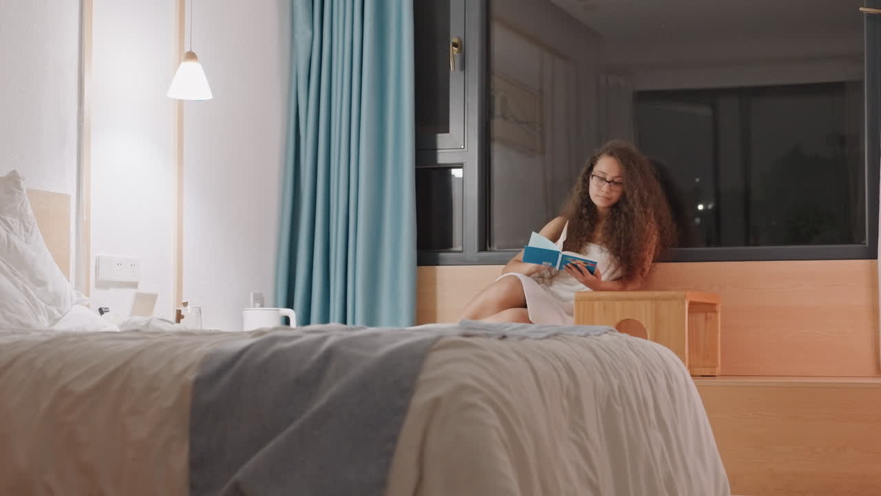 Relaxing night scene. Comfortable bed setting with woman enjoying evening reading activity. Tranquil hotel environment featuring woman reading by window beneath gentle lamp illumination